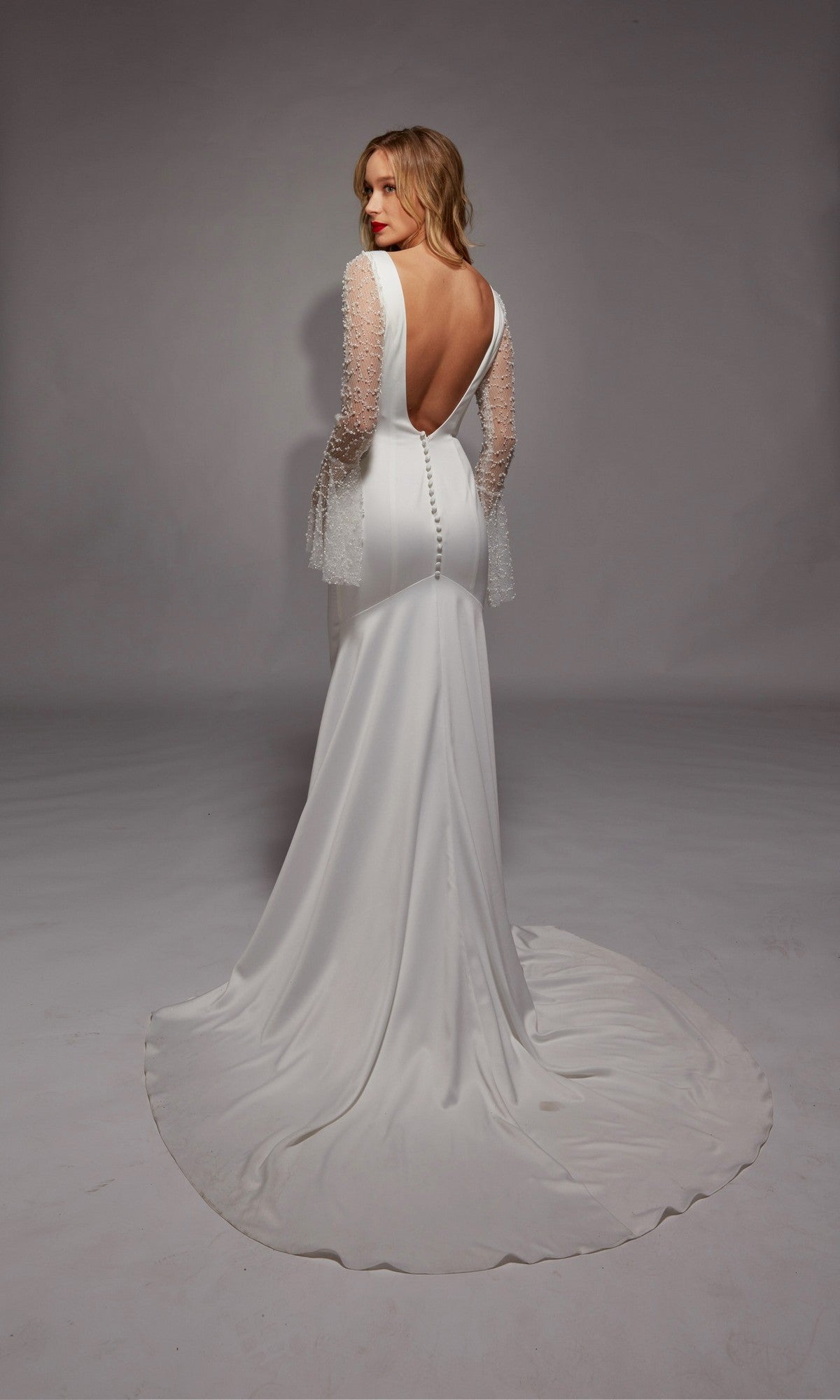 A woman in the Long White Dress: Alyce 7137, featuring illusion lace sleeves and an open back, stands on a plain background with her back to the camera.